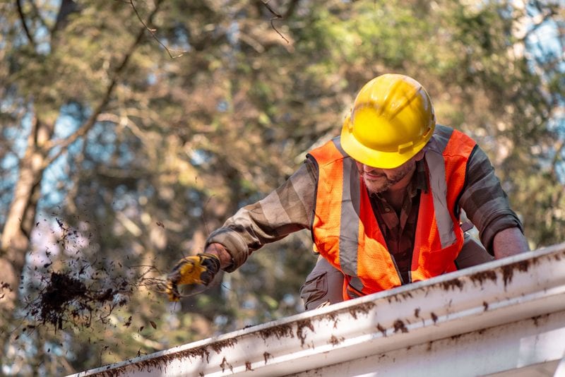 A worker on a ladder clearing leaves out of a roof gutter.